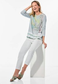 Langarmoberteil in Hellblau mit einem bunten abstrakten Muster. Weiße Slim-Fit-Hosen und grüne strukturierte Sandalen. Model sitzt auf einem weißen Block.