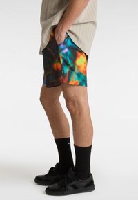 Bunte Badeshorts mit einem Batikmuster in Orange-, Blau- und Grüntönen. Sie verfügen über Seitentaschen und werden mit schwarzen Schuhen und Socken kombiniert.