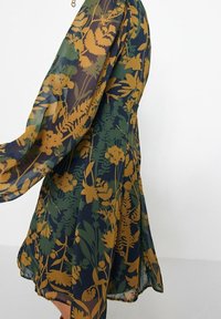 Šaty s květinovým potiskem z průsvitné látky, s tmavě modrým podkladem a hořčicovými botanickými vzory. Dlouhé, splývavé rukávy a rozšířený střih.