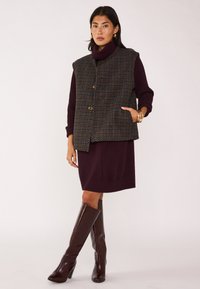 Gilet in misto lana con motivo a quadri, dotato di colletto alto, bottoni frontali e tasche laterali, indossato sopra un vestito bordeaux e stivali al ginocchio.