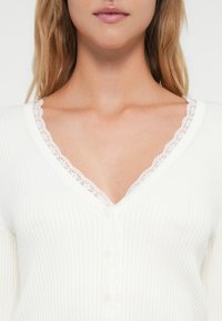 Κοντινό πλάνο μιας γυναίκας που φοράει ένα λευκό ριμπ βελονάκι με V-λαιμό, πάνω από ένα λευκό τοπ με δαντέλα, αποκαλύπτοντας τους ώμους και το άνω στήθος.