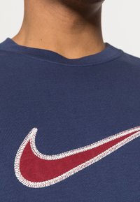 Tmavě modré bavlněné tričko s velkým červeným logem Nike ve tvaru háčku s bílými šicími akcenty. Kulatý výstřih a žebrovaná textura.