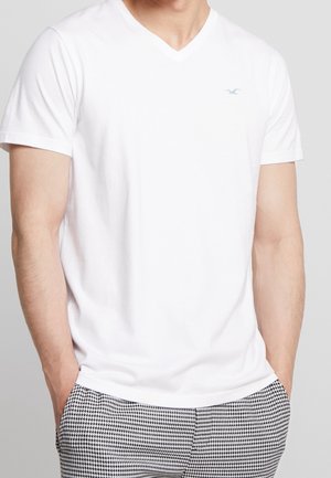 Homme portant un t-shirt blanc uni à col en V avec un petit logo bleu et un pantalon à motif pied-de-poule noir et blanc, mains dans les poches.