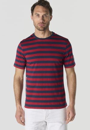 Hombre con camiseta de manga corta a rayas horizontales en azul marino y rojo y pantalones blancos, de pie contra un fondo claro y liso.