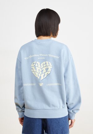 Osoba o krótkich ciemnych włosach nosi jasnoniebieską bluzę z graficznym napisem w kształcie serca „CATCH ART WIP” na plecach, w połączeniu z niebieskimi szortami dżinsowymi.