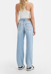 Femme portant un jean large bleu clair, un débardeur blanc et des baskets noires et blanches, debout de dos sur un fond blanc.