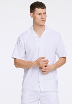 Camisa blanca de manga corta y botón, con cuello, hecha de un tejido suave y texturizado. Presenta un bolsillo en el pecho y un corte holgado.