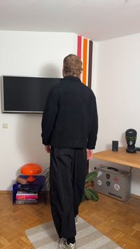 Juoda vilnos striukė su aukštu apykakliu, derinama su laisvomis juodomis kelnėmis. Kambarys pasižymi ryškiomis sienų juostomis ir oranžine lempa.