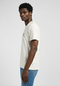 Camiseta de algodón de color beige claro, con mangas cortas y cuello redondo. Cuenta con un pequeño gráfico en el pecho y un corte relajado. Superficie texturizada.