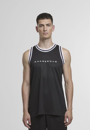 Młody mężczyzna ubrany w czarną sportową koszulkę bez rękawów z białą lamówką i napisem "DANGEROUS", stojący na tle jednolitego szarego tła.