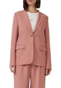 Vrouw in een stofroze getailleerde blazer met één knop, bijpassende broek en een wit T-shirt met kleine zwarte tekst.