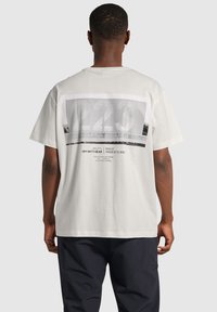 Vit bomullströja med en stor grafisk design på baksidan som har nummer "223" och textinslag. Kort ärm, avslappnad passform.