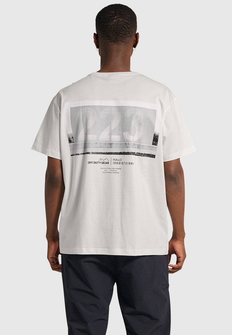 Vit bomullströja med en stor grafisk design på baksidan som har nummer "223" och textinslag. Kort ärm, avslappnad passform.