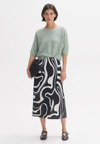 Lichtgroene gebreide trui met korte mouwen, gecombineerd met een zwart-witte rok met golvenpatroon. Model houdt een zwarte gewatteerde clutch vast.