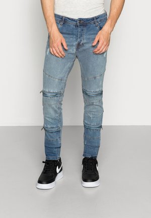 Homme portant un jean denim bleu bicolore avec plusieurs fermetures éclair horizontales au-dessus des genoux et des baskets noires avec des semelles blanches.