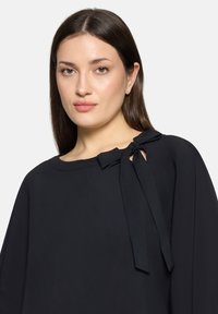 Zwarte blouse met een losse pasvorm, ronde halslijn en lintdetail bij de schouder. Gladde stof met minimale textuur.