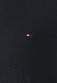 Pull en tricot noir avec un petit logo tricolore aux accents rouge, blanc et bleu sur le côté gauche. Texture douce, coupe classique.