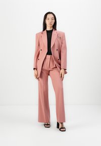 Růžový dvouřadý blazer a odpovídající široké kalhoty, kombinované s černým texturovaným topem. Obsahuje malou sponu ve tvaru mašle na blazeru.