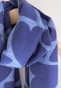 Sciarpa di lana con motivo a cuori blu e navy, dalla texture morbida e aspetto drappeggiato, appesa a una gruccia di legno.