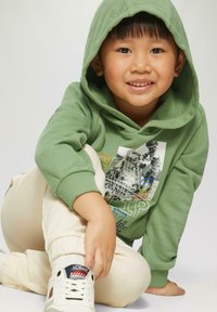 Zielona bluza z kapturem z graficznym nadrukiem, kremowe dresy i białe sneakersy z czerwonymi i niebieskimi akcentami. Dziecko siedzi i się uśmiecha.