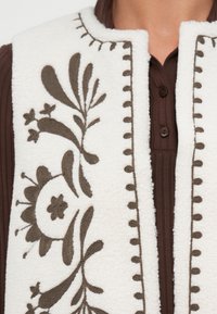 Gilet en polaire blanc avec broderie florale marron. Il présente un col côtelé marron avec boutons et des coutures décoratives le long des bords.
