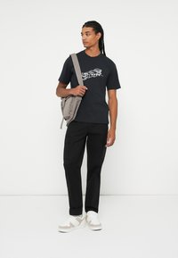 Tricou negru din bumbac cu un design grafic, purtat împreună cu pantaloni cargo negri și adidași albi cu accente bej. Modelul poartă o geantă de umăr gri.