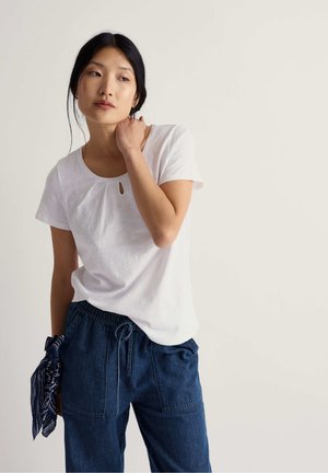 Camiseta blanca de manga corta con cuello redondo y un pequeño detalle de abertura en forma de ojo de cerradura, combinada con pantalones azul oscuro con cordón y un accesorio con estampado.