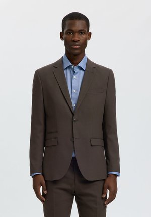Homme portant une veste de costume marron foncé sur une chemise bleu clair boutonnée, debout devant un fond clair uni, regardant devant.