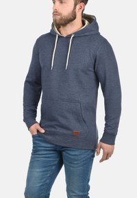 Sudadera con capucha en tejido azul oscuro, con un bolsillo delantero, cordones crema y un acabado texturizado con una pequeña etiqueta de cuero.