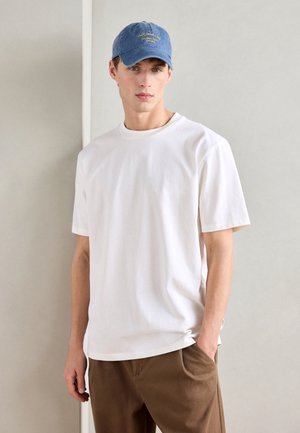 White cotton t-shirt with short sleeves, paired with brown trousers. A blue cap with embroidered text sits on the model's head.