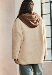 Sweatshirt in pile beige con cappuccio marrone, vestibilità ampia, maniche lunghe, superficie strutturata e stile casual e rilassato.