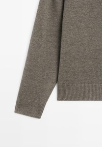 Šedý pletený svetr s žebrovaným límcem a manžetami, který má měkkou texturu a mírně volný střih. Bez viditelných vzorů nebo kovových detailů.