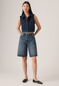 Ensemble en jean comprenant un gilet et un short en denim bleu délavé. Le gilet possède des poches frontales et une fermeture à boutons ; le short a une taille mi-haute et une coupe droite. Des ballerines noires.