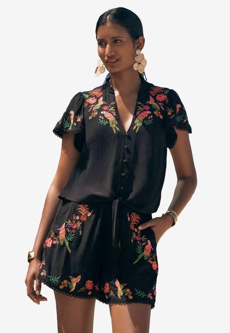 Black embroidered romper with short sleeves, featuring colorful floral and bird patterns, button detail, and a tie at the waist.