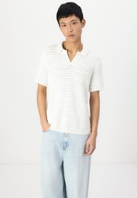 Polo blanco de punto con manga corta y cuello en V, con un diseño texturizado a rayas. Combinado con unos pantalones vaqueros de pierna ancha en azul claro.