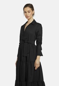 Schwarz gestuftes Kleid mit Kragen, langen Ärmeln und einem Stoffgürtel. Glatte Textur und fließendes Design mit gerafften Details.