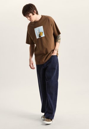 Camiseta de algodón marrón con un estampado gráfico que muestra un fantasma y una bebida, combinada con jeans de mezclilla oscuros y de corte ancho, y zapatillas con estampado de leopardo.