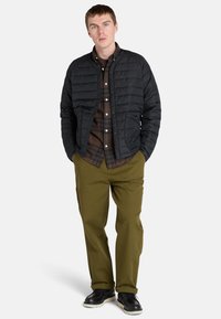 Zwarte gewatteerde jas met een opstaand kraag, gecombineerd met een bruine geruite shirt en olijfgroene broek. Zwarte schoenen maken de outfit compleet.
