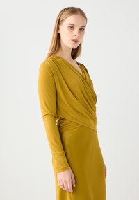 Langarmige jurk van mosterdgele stof, met een gedrapeerde V-halslijn en een gegatherde taille, die een vloeiende silhoutte creëert.