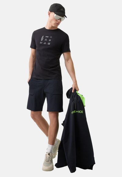 Sort t-shirt med grafiske symboler, navy shorts og en mørk hoodie med neonlys. Modellen bærer spejlede solbriller og lyse sneakers.