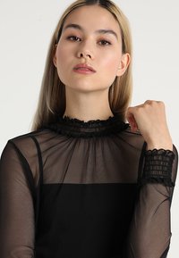 Čierny priesvitný top s vysokým volánovým golierom a elastickými krajkovými manžetami. Hladká textúra s priliehavým strihom. Dlhé rukávy, minimalistický vzor.