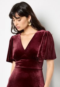 Burgundské sametové šaty s výstřihem do V, krátkými balónovými rukávy a nařaseným detailem v pase. Zlaté kruhové náušnice doplňují outfit.