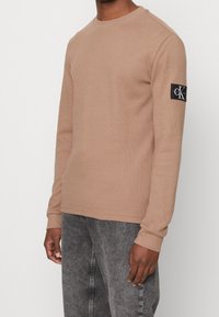 Hellbrauner gestrickter Langarmpullover mit Rundhalsausschnitt und gerippten Bündchen, ausgestattet mit einem schwarzen Logopatch am linken Ärmel.