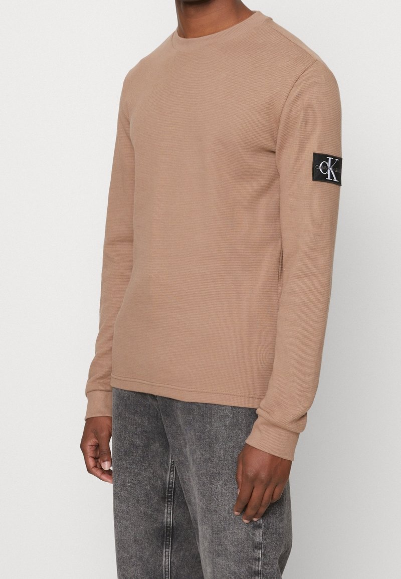 Hellbrauner gestrickter Langarmpullover mit Rundhalsausschnitt und gerippten Bündchen, ausgestattet mit einem schwarzen Logopatch am linken Ärmel.