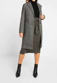 Abrigo gris de espiga con ajuste entallado, solapas con muesca y dos bolsillos delanteros, combinado con una falda verde oscuro y botines negros.