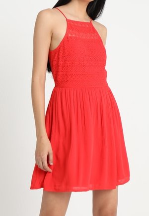 Mujer vestida con un vestido rojo sin mangas con un cuerpo de encaje y una falda plisada, posando frente a un fondo blanco liso.