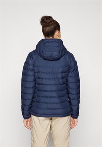 Marineblå dunjakke med hette, med horisontal quiltet design, glatt tekstur og elastiske mansjetter. Båret over beige cargo bukser.