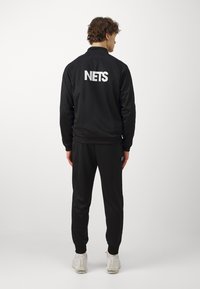 Tuta nera con giacca a zip e pantaloni, con la scritta "NETS" stampata in bianco sul retro. Texture liscia, polsini e vita a coste.