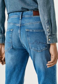 Man die lichtblauwe spijkerbroek en een bijpassend spijkerjasje met geknoopte manchetten draagt, van achteren weergegeven bij de taille en heupen.