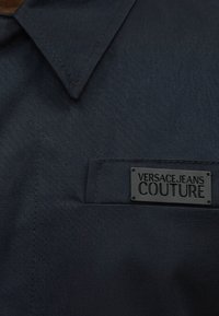 Σκούρο μπλε ύφασμα με μυτερό γιακά και τσεπάκι στο στήθος με μαύρη ορθογώνια ετικέτα Versace Jeans Couture.